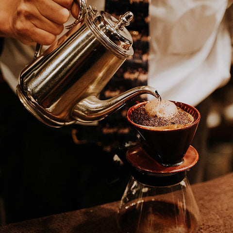 Pour over coffee brewing with a stainless steel gooseneck kettle, ceramic dripper, and glass server, creating fresh specialty coffee for home baristas.